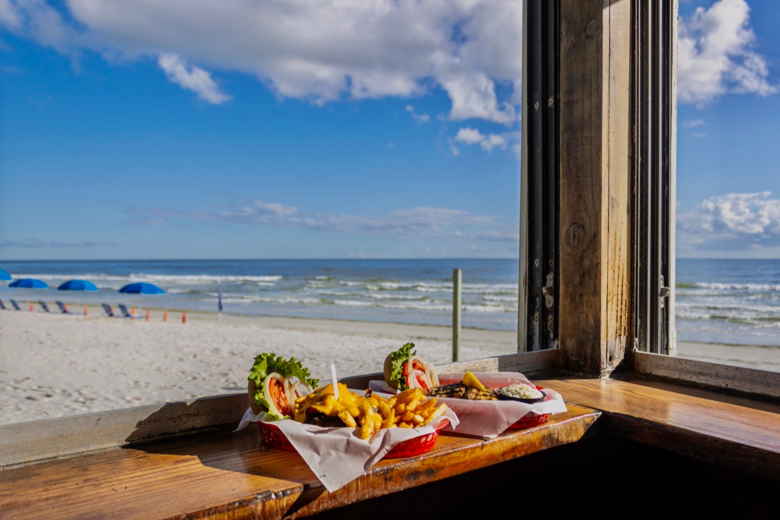 A burger and fries basket beside a grilled mahi plate, with the beach visible through the window