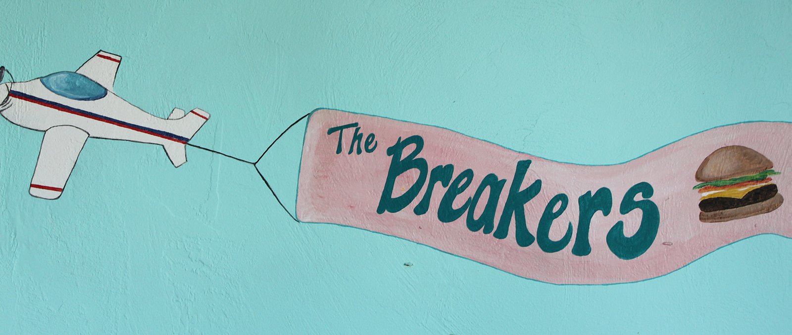 Hand-painted mural of a plane pulling a pink banner that reads The Breakers with a burger on it